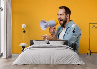 Crazy young man in casual blue shirt posing isolated on yellow orange wall background studio portrait. People lifestyle concept. Mock up copy space. Screaming in megaphone pointing index finger aside. Wall mural