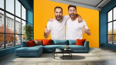 Crazy young men guys friends in white blank empty t-shirts posing isolated on yellow orange wall background studio portrait. People lifestyle concept. Mock up copy space. Screaming, showing thumbs up. Wall mural