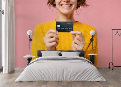 Cropped image of smiling young brunette woman girl in yellow sweater posing isolated on pastel pink wall background studio portait. People lifestyle concept. Mock up copy space. Hold credit bank card. Wall mural