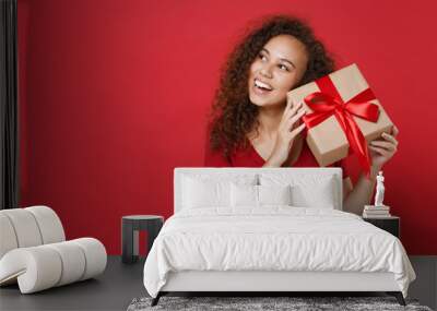 Curious funny young african american woman girl in casual t-shirt posing isolated on red background. St. Valentine's Day, Women's Day, birthday, holiday concept. Hold present box with gift ribbon bow. Wall mural