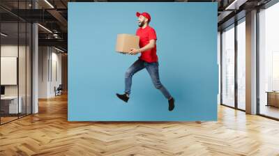 Delivery man in red uniform isolated on blue background, studio portrait. Male employee in cap t-shirt print working as courier dealer hold empty cardboard box. Service concept. Mock up copy space. Wall mural