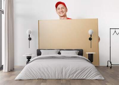 Delivery man in red uniform isolated on white background. Male in cap, t-shirt, jeans working as courier or dealer holding big large heavy empty blank cardboard box. Receiving package. Copy space area Wall mural