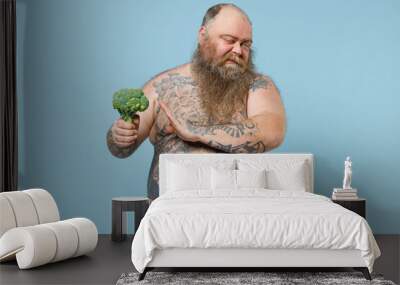 Disgusted fat pudge obese chubby overweight man has tattooed big belly showing stop gesture with palm to fresh vegetable broccoli isolated on blue background. Weight loss obesity healthy diet concept. Wall mural