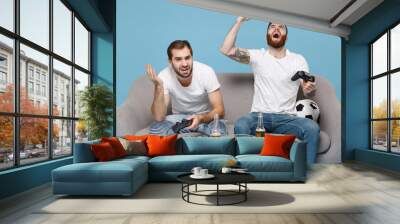 Displeased crazy guys friends sit on couch isolated on blue background. Sport leisure concept. Cheer up support favorite team with soccer ball play game with joystick screaming doing winner gesture. Wall mural