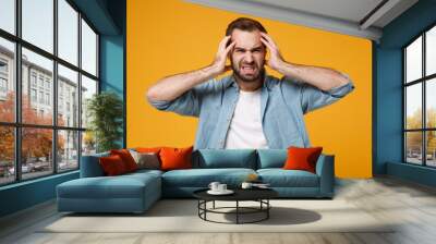 Displeased exhausted young bearded man in casual blue shirt posing isolated on yellow orange background, studio portrait. People emotions lifestyle concept. Mock up copy space. Putting hands on head. Wall mural