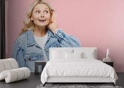 Elderly curious nosy happy woman 50s wearing denim jacket try to hear you overhear listening intently isolated on plain pastel light pink background studio portrait. People lifestyle fashion concept. Wall mural