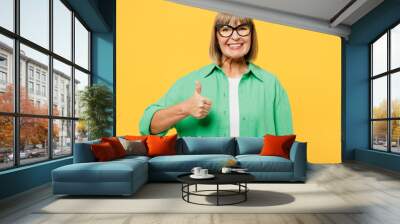 Elderly smiling happy cheerful blonde woman 50s years old wear green shirt glasses casual clothes showing thumb up like gesture isolated on plain yellow background studio portrait. Lifestyle concept. Wall mural