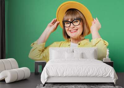 Elderly smiling happy fashionable woman 50s wearing glasses yellow shirt white t-shirt touch hat looking camera isolated on plain green background studio portrait People emotions lifestyle concept. Wall mural