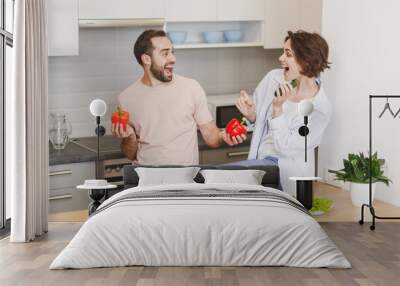 Excited couple friends guy girl sit on table preparing vegetable salad cooking food in light kitchen at home. Dieting family healthy lifestyle concept. Mock up copy space. Hold cucumber bell peppers. Wall mural