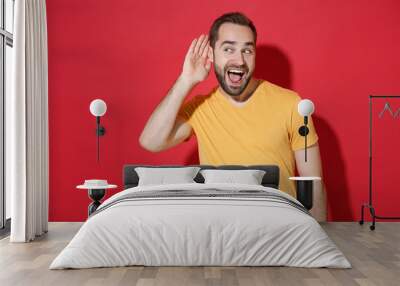 Excited curious young bearded man guy in casual yellow t-shirt posing isolated on red wall background studio portrait. People lifestyle concept. Mock up copy space. Try to hear you listening intently. Wall mural