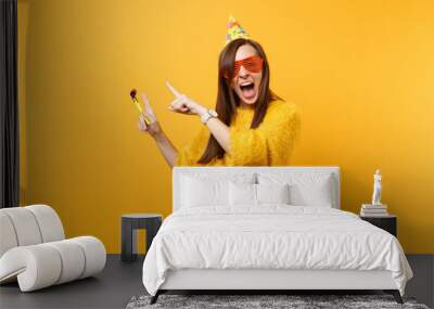 Excited happy woman in orange funny glasses, birthday hat with playing pipe pointing index fingers aside on copy space, celebrating isolated on yellow background. People sincere emotions, lifestyle. Wall mural