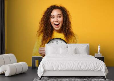 Excited young african american girl in fur sweater posing isolated on yellow orange background, studio portrait. People lifestyle concept. Mock up copy space. Holding clock, keeping mouth wide open. Wall mural