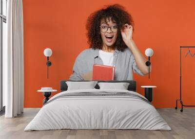 Excited young african american student girl in casual clothes, eyeglasses isolated on orange background studio portrait. People lifestyle concept. Mock up copy space. Hold books, keeping mouth open. Wall mural