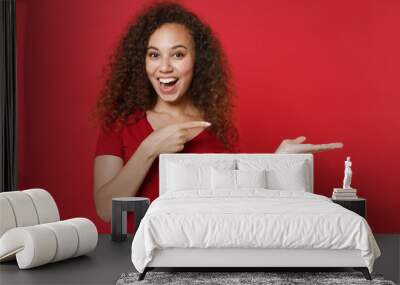 Excited young african american woman girl in casual t-shirt posing isolated on red wall background studio portrait. People lifestyle concept. Mock up copy space. Pointing index finger hand aside. Wall mural