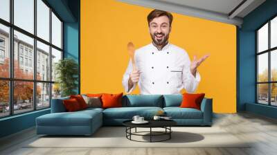 Excited young bearded male chef cook or baker man in white uniform shirt posing isolated on yellow background in studio. Cooking food concept. Mock up copy space. Hold wooden spoon, spreading hands. Wall mural