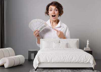 Excited young business woman in white shirt isolated on grey background. Achievement career wealth business concept. Mock up copy space. Pointing index finger on fan of cash money in dollar banknotes. Wall mural