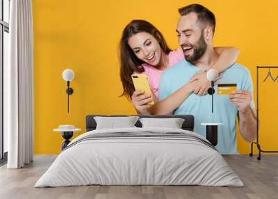 Excited young couple two friends guy girl in blue pink t-shirts isolated on yellow background studio. People lifestyle concept. Mock up copy space. Using mobile phone, hold credit bank card, hugging. Wall mural