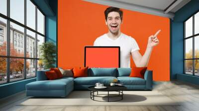 Excited young man 20s wearing basic casual white t-shirt hold laptop pc computer with blank empty screen pointing index finger aside up isolated on bright orange colour background studio portrait. Wall mural