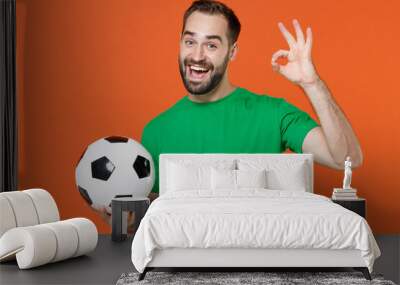 Excited young man football fan in green t-shirt cheer up support favorite team with soccer ball showing OK sign isolated on orange background studio portrait. People sport leisure lifestyle concept. Wall mural