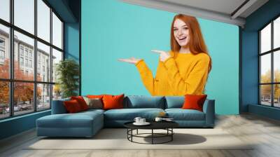 Excited young redhead woman girl in yellow sweater posing isolated on blue turquoise wall background studio portrait. People lifestyle concept. Mock up copy space. Pointing index finger, hand aside. Wall mural