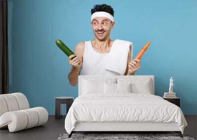 Excited young sporty fitness man with thin skinny body sportsman in white headband shirt shorts hold two vegetables carrot cucumber isolated on blue background. Workout gym sport motivation concept. Wall mural