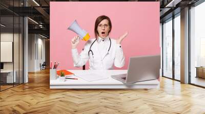 Female doctor sit at desk work on computer with medical document hold megaphone in hospital isolated on pastel pink background. Woman in medical gown glasses stethoscope. Healthcare medicine concept. Wall mural