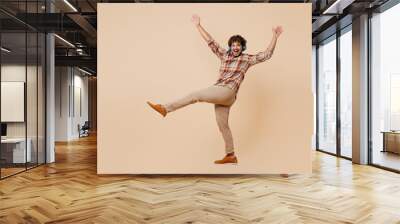 Full body fun young Indian man wear brown shirt casual clothes headphones listen to music dance on party gesticulating hands isolated on plain pastel light beige background. People lifestyle concept. Wall mural