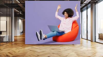 Full body fun young woman of African American ethnicity 20s in pink striped shirt sit in bag chair hold use work on laptop pc computer raise up hands isolated on plain pastel light purple background Wall mural