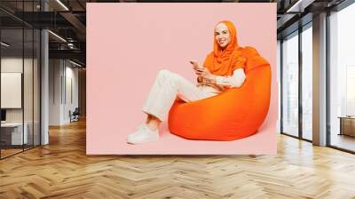 Full body happy fun young arabian asian muslim woman wears orange abaya hijab sit in bag chair hold use mobile cell phone isolated on plain pink background. Uae middle eastern islam religious concept. Wall mural