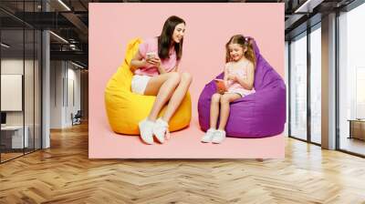 Full body happy woman wear casual clothes with child kid girl 6-7 years old. Mother daughter sit in bag chair hold use mobile cell phone isolated on plain pink background. Family parent day concept. Wall mural