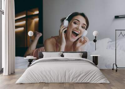 Full body housewife excited young woman wear pajamas headphones lying in bed listen music dancing enjoy new playlist rest relax on weekends indoors at home. Good mood night morning bedtime concept Wall mural