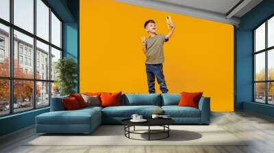 Full body little small happy boy 6-7 years old in green t-shirt do selfie shot on mobile cell phone show v-sign gesture isolated on plain yellow background. Mother's Day love family lifestyle concept. Wall mural