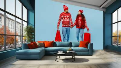 Full body merry young couple two man woman in red Christmas sweater Santa hat posing hat hold hand shopping package bag walk go isolated on plain pastel blue background Black Friday sale buy concept Wall mural