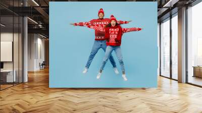 Full body merry young couple two man woman wear red Christmas sweater Santa hat posing jump high with outstretched hands isolated on plain pastel blue background Happy New Year 2023 holiday concept. Wall mural