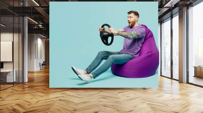 Full body shocked excited fun young man he wears purple t-shirt hold steering wheel driving car sit in bag chair isolated on plain pastel light blue cyan background studio portrait. Lifestyle concept. Wall mural