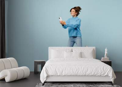 Full body side view happy young woman wear knitted sweater jump high hold in hand use mobile cell phone isolated on plain pastel light blue cyan background studio portrait. People lifestyle concept. Wall mural