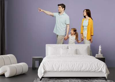 Full body side view young parents mom dad child kid daughter girl 6 years old wearing blue yellow casual clothes hold hands walk go point aside isolated on plain purple background. Family day concept. Wall mural