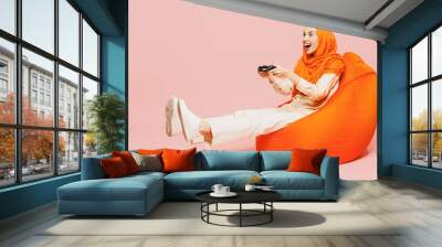 Full body young arabian asian muslim woman wear orange abaya hijab sit in bag chair hold in hand play pc game with joystick console isolated on plain pink background. Uae middle eastern islam concept. Wall mural