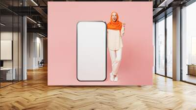 Full body young arabian muslim woman wear orange abaya hijab big huge blank screen mobile cell phone with area point index finger up isolated on plain pink background Uae middle eastern islam concept Wall mural