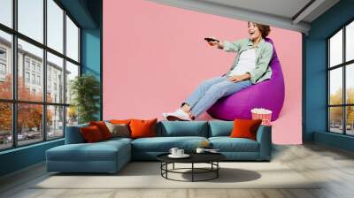 Full body young excited fun woman she wear green shirt white t-shirt sit in bag chair hold popcorn bucket watch TV show movie switch channels with remote isolated on plain pastel light pink background Wall mural