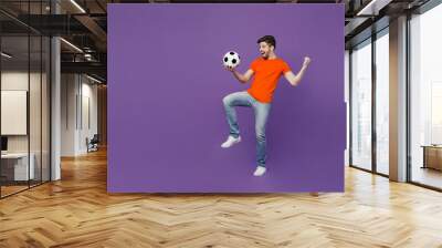 Full body young fan man he wear orange t-shirt cheer up support football sport team hold juggling on knee soccer ball watch tv live stream sit in bag chair isolated on plain dark purple background Wall mural