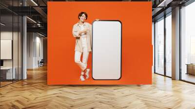 Full body young happy caucasian woman she wear beige shirt casual clothes point finger on big huge blank screen mobile cell phone smartphone with area isolated on plain orange red background studio. Wall mural