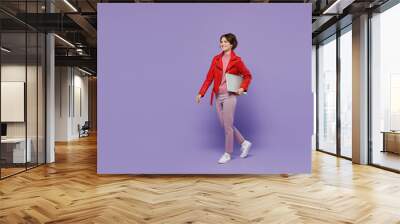 Full body young happy fun woman 20s in red leather jacket hold closed laptop pc computer walk going isolated on plain pastel light purple background studio portrait. People lifestyle fashion concept Wall mural