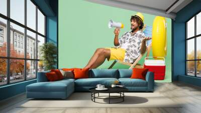 Full body young happy Indian man he wear beach shirt hat rest near hotel pool sit in deckchair scream shout in megaphone isolated on plain green color background. Summer vacation sea sun tan concept. Wall mural