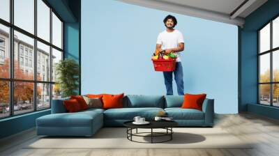 Full body young happy Indian man wear white t-shirt casual clothes hold red basket for takeaway mock up with food products isolated on plain blue background. Delivery service from shop or restaurant. Wall mural