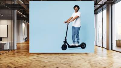 Full body young happy smiling Indian man he wear white t-shirt casual clothes ride electric scooter look camera isolated on plain pastel light blue cyan background studio portrait. Lifestyle concept. Wall mural