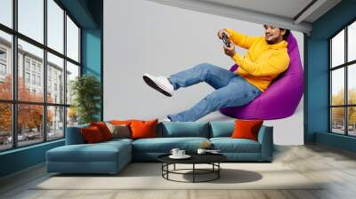 Full body young Indian man he wearing casual yellow hoody sit in bag chair hold in hand play pc game with joystick console isolated on plain grey background studio portrait. People lifestyle portrait. Wall mural