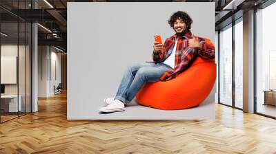 Full body young Indian man wear red shirt casual clothes sit in bag chair hold in hand use point finger on mobile cell phone isolated on plain grey color background studio portrait. Lifestyle concept Wall mural