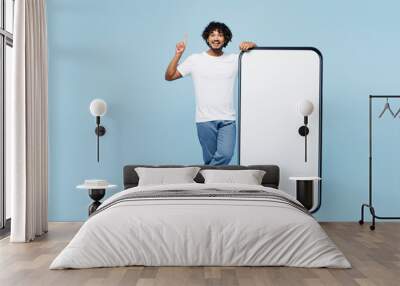 Full body young Indian man wear white t-shirt casual clothes big huge blank screen mobile cell phone smartphone with area point finger up isolated on plain pastel blue background. Lifestyle concept. Wall mural