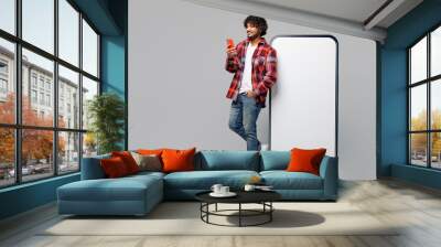 Full body young Indian man wears red shirt casual clothes stand near big huge blank screen mobile cell phone with workspace copy space area use hold smartphone isolated on plain grey color background. Wall mural